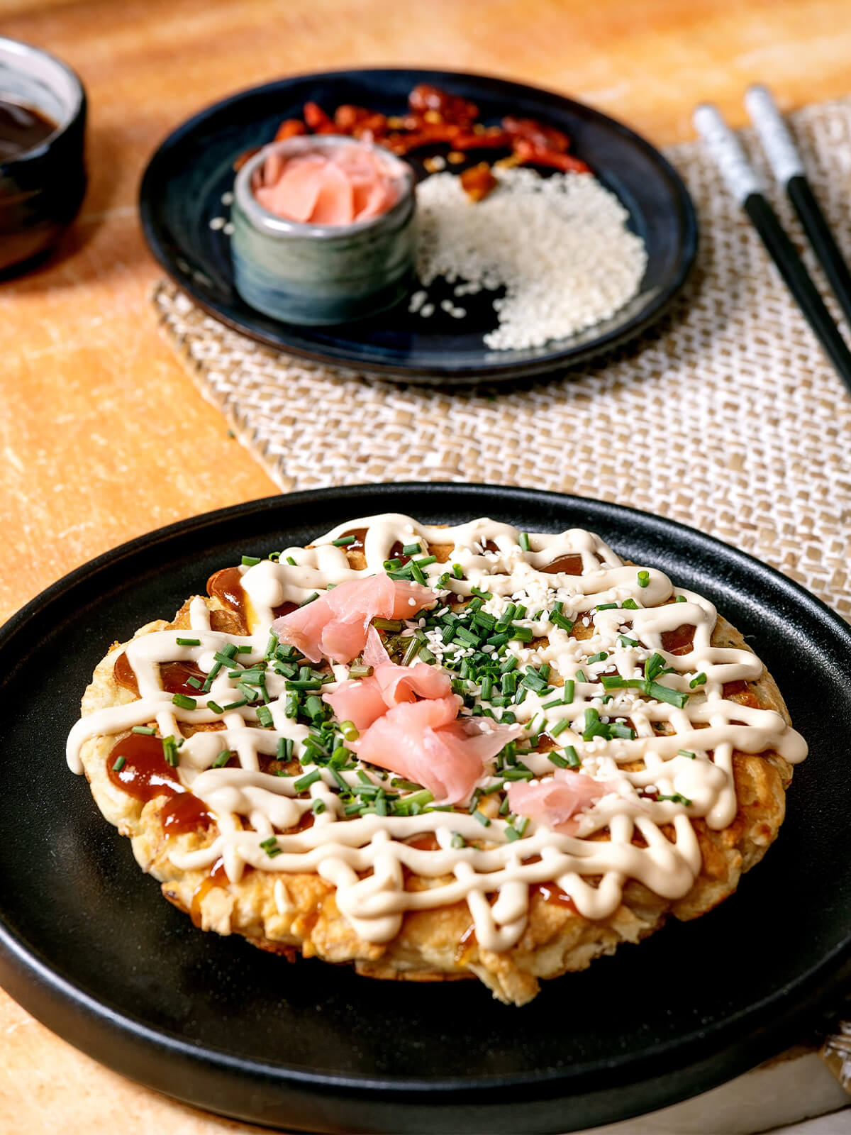 Okonomiyaki, japanischer Kohl-Pfannkuchen, mit Mayonnaise-Gitter, Okonomiyaki-Sauce, rosa eingelegtem Ingwer, Schnittlauch und Sesam auf schwarzem Teller