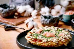Okonomiyaki auf einem schwarzen Teller, mit Mayonnaise-Streifen, dunkler Sauce, rosa eingelegtem Ingwer und Schnittlauch garniert