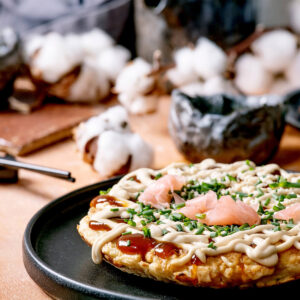 Okonomiyaki auf einem schwarzen Teller, mit Mayonnaise-Streifen, dunkler Sauce, rosa eingelegtem Ingwer und Schnittlauch garniert