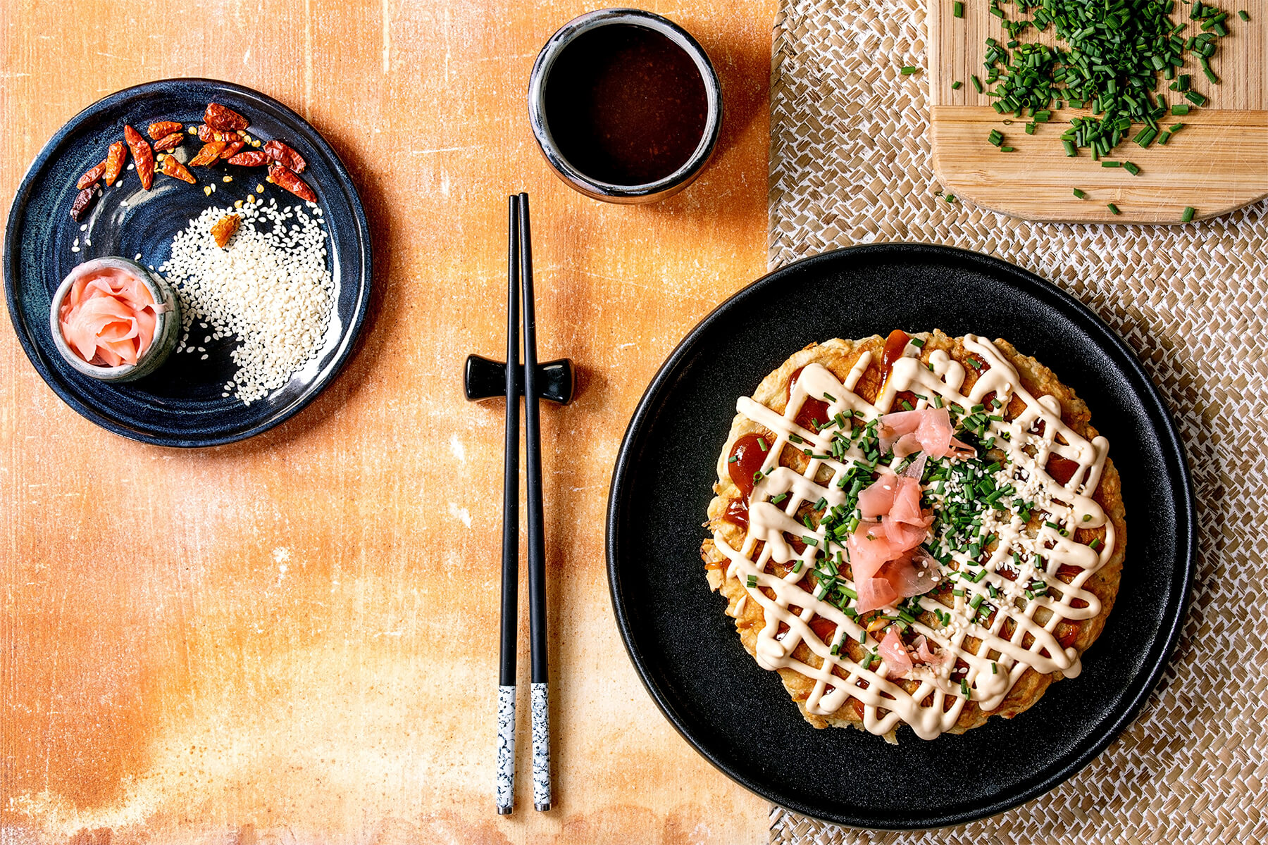 Okonomiyaki von oben auf schwarzem Teller, daneben Stäbchen, eine Schale Okonomiyaki-Sauce und ein Tellerchen mit Sesam und eingelegtem Ingwer