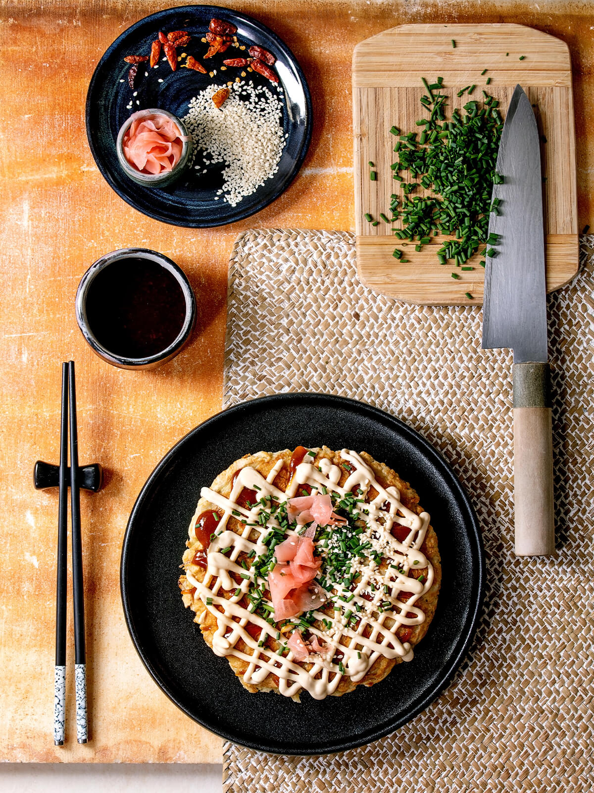 Okonomiyaki von oben auf schwarzem Teller, daneben ein japanisches Messer mit fein geschnittenem Schnittlauch und Sesam