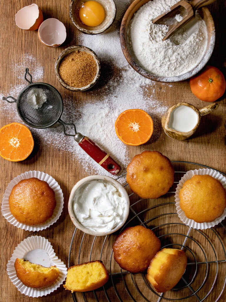 Zutaten für Orangen-Muffins: Mehl, Eier, brauner Zucker, Orangen, Milch und fertige Muffins auf Holz