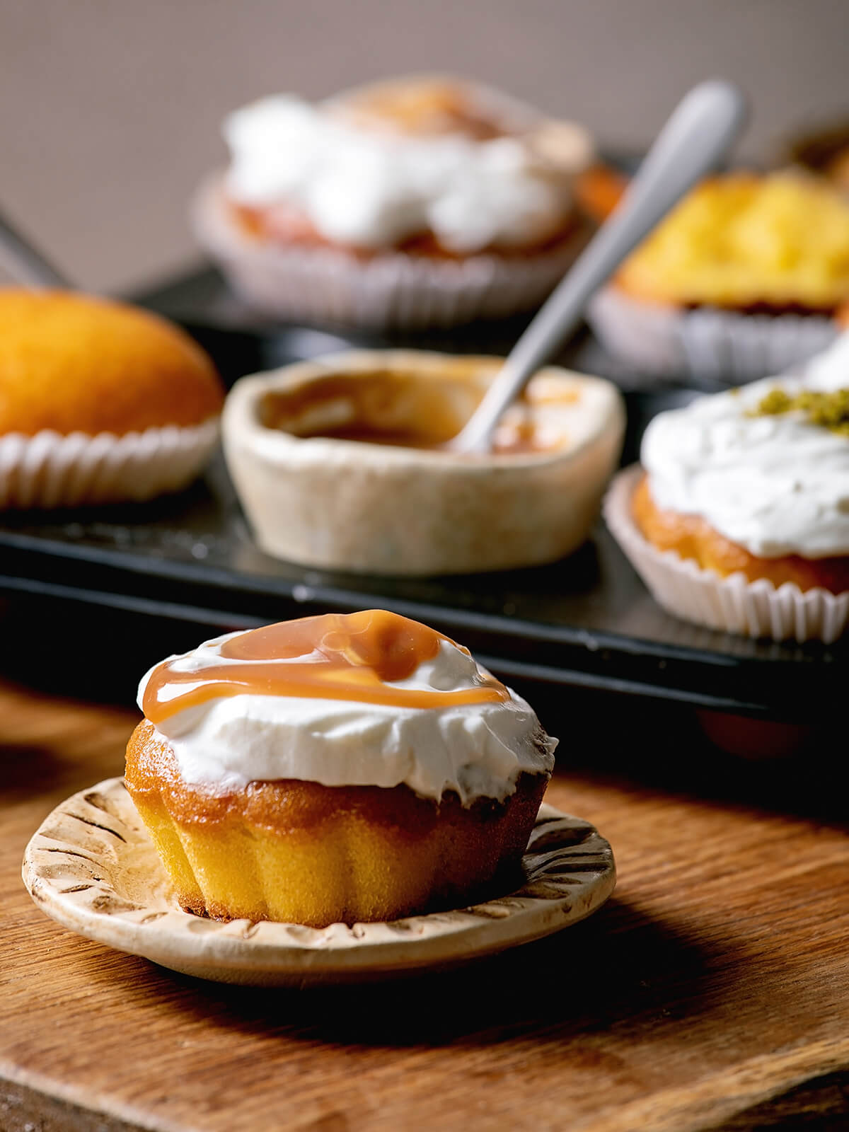 Orangen-Muffin mit Sahnehaube und Karamell-Swirl auf einem kleinen Teller, dahinter weitere Muffins und eine Schale Karamell
