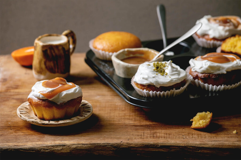 Mehrere Orangen-Muffins mit Sahnehaube und Karamell auf einem dunklen Tablett, daneben eine Schale mit Karamellsauce