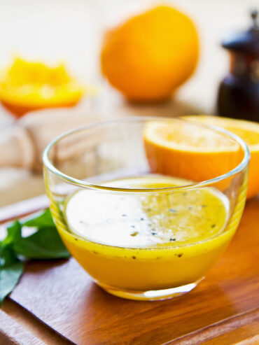 Goldorange Orangen-Vinaigrette mit schwarzem Sesam in einer Glasschale, daneben halbierte Orangen und Basilikum