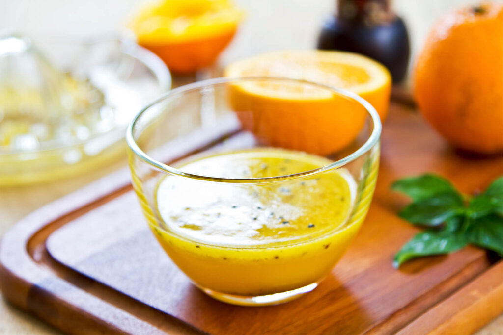 Orangen-Vinaigrette mit schwarzem Sesam in einer Glasschale, daneben eine ausgepresste Orange und eine Zitruspresse