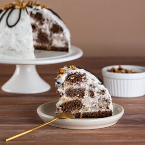Weiße Pancho-Kuppeltorte auf einem Tortenständer mit Schokoladen-Schlaufen und Walnuss-Krone, davor ein aufgeschnittenes Stück mit sichtbarem Mosaik aus Schokoladenbiskuit, heller Creme und Ananasstücken