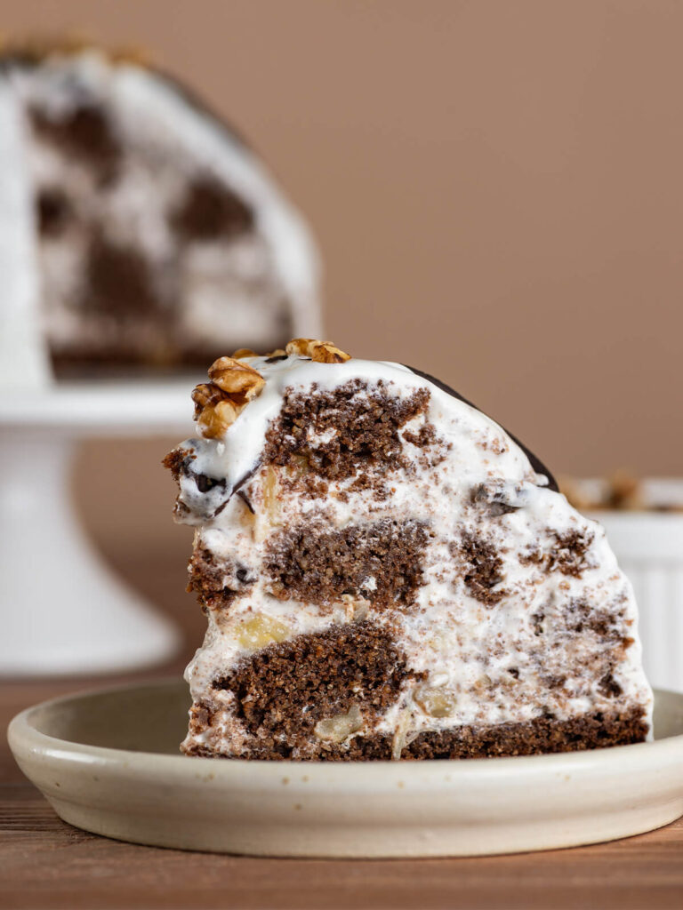 Aufgeschnittenes Stück Pancho-Torte auf einem Teller in Nahaufnahme, im Anschnitt das Mosaik aus dunklem Schokoladenbiskuit, weißer Sauerrahm-Creme, hellen Ananasstücken und Walnüssen, oben ein Schokoladen-Bogen und eine Walnuss