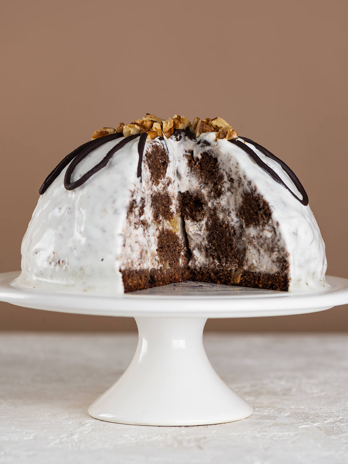 Angeschnittene Pancho-Kuppeltorte auf einem weißen Tortenständer, im Anschnitt das Mosaik aus dunklem Schokoladenbiskuit, heller Creme und Ananasstücken, oben Schokoladen-Schlaufen und Walnüsse