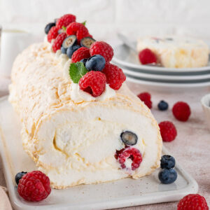 Pavlova-Roulade auf einem weißen Keramikbrett, die knusprige Baiser-Hülle im Anschnitt mit Sahne und frischen Himbeeren und Heidelbeeren, obenauf Sahne-Tupfen, Beeren und Minze