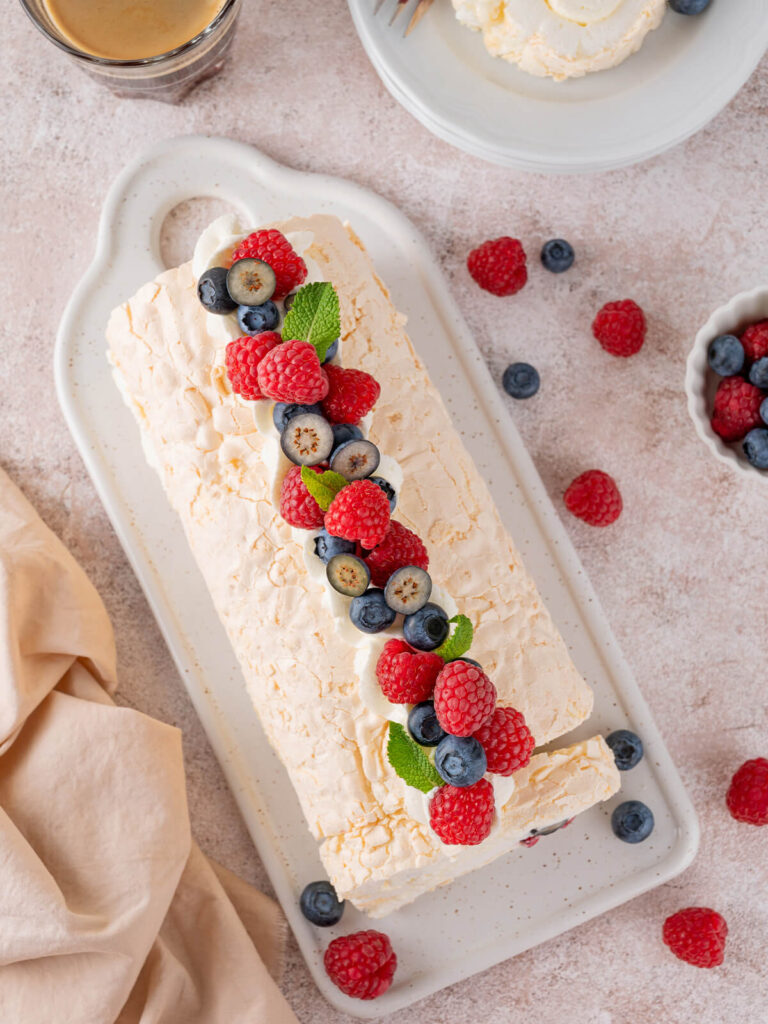Fertige Pavlova-Roulade von oben auf einem weißen Keramikbrett mit Griff, dekoriert mit einer Reihe frischer Himbeeren, ganzer und halbierter Heidelbeeren, Minzblättern und Sahne-Tupfen