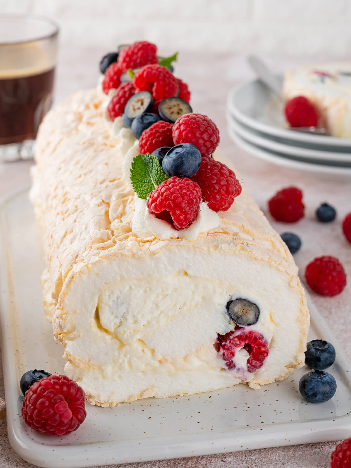 Pavlova-Roulade in Nahaufnahme auf einem weißen Keramikbrett, die knusprige Baiser-Hülle im Anschnitt mit Sahne und frischen Himbeeren und Heidelbeeren, obenauf Sahne-Tupfen, Beeren und Minze