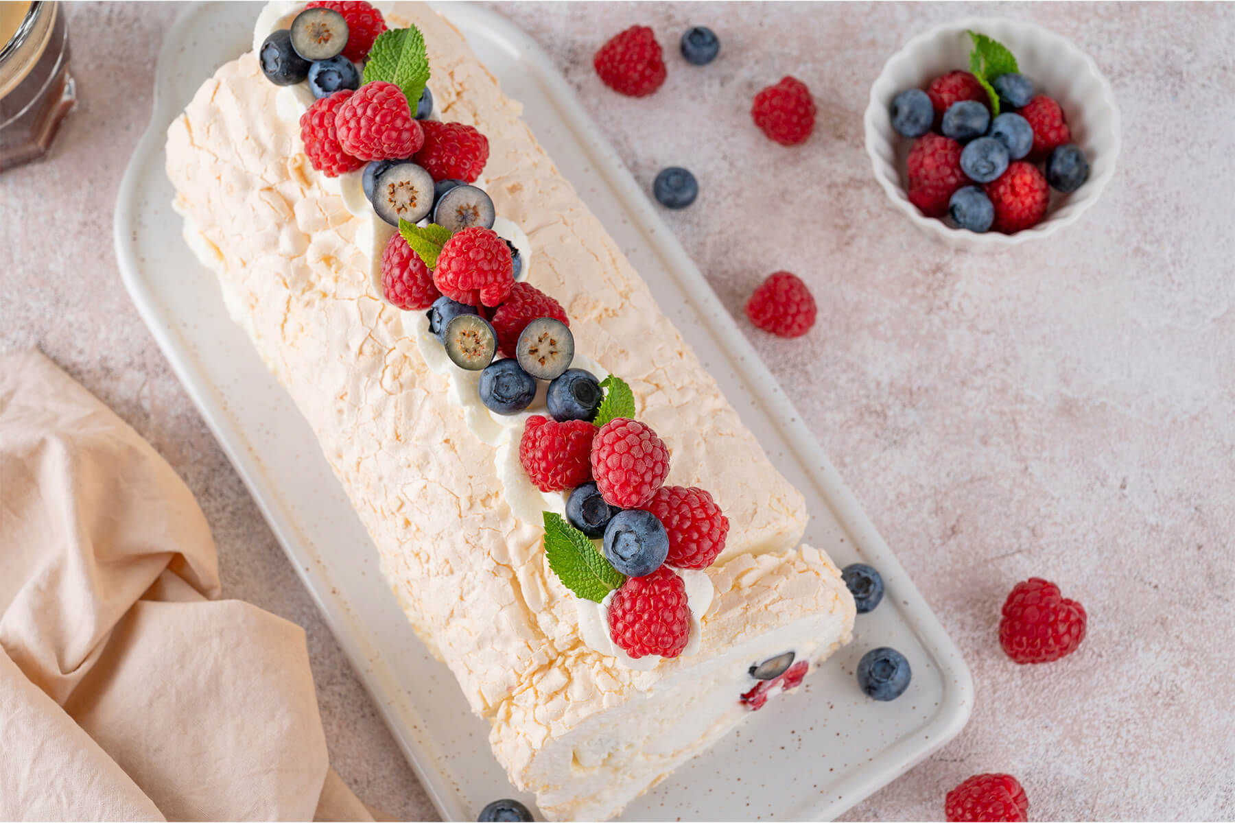 Pavlova-Roulade von oben auf einem weißen Keramikbrett, mit einer Reihe aus frischen Himbeeren, ganzen und halbierten Heidelbeeren, Minzblättern und Sahne-Tupfen verziert, daneben ein Schälchen Beeren