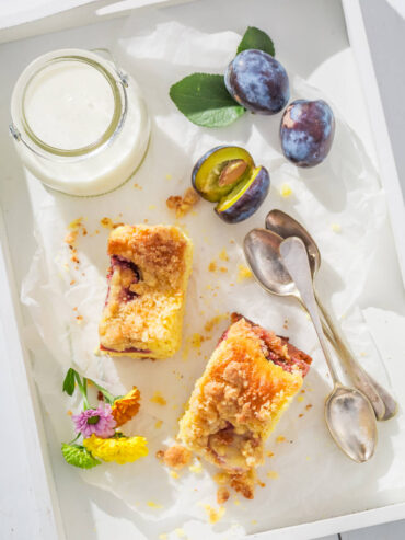 Zwei hohe Stücke Pflaumen-Streuselkuchen mit dicker Streuselkrone und Pflaumen im Teig auf einem hellen Tablett, daneben frische Zwetschgen