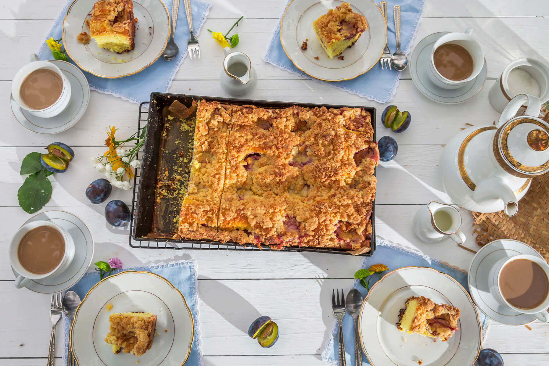 Pflaumen-Streuselkuchen von oben auf einer gedeckten Kaffeetafel mit Tassen, Kannen und frischen Pflaumen