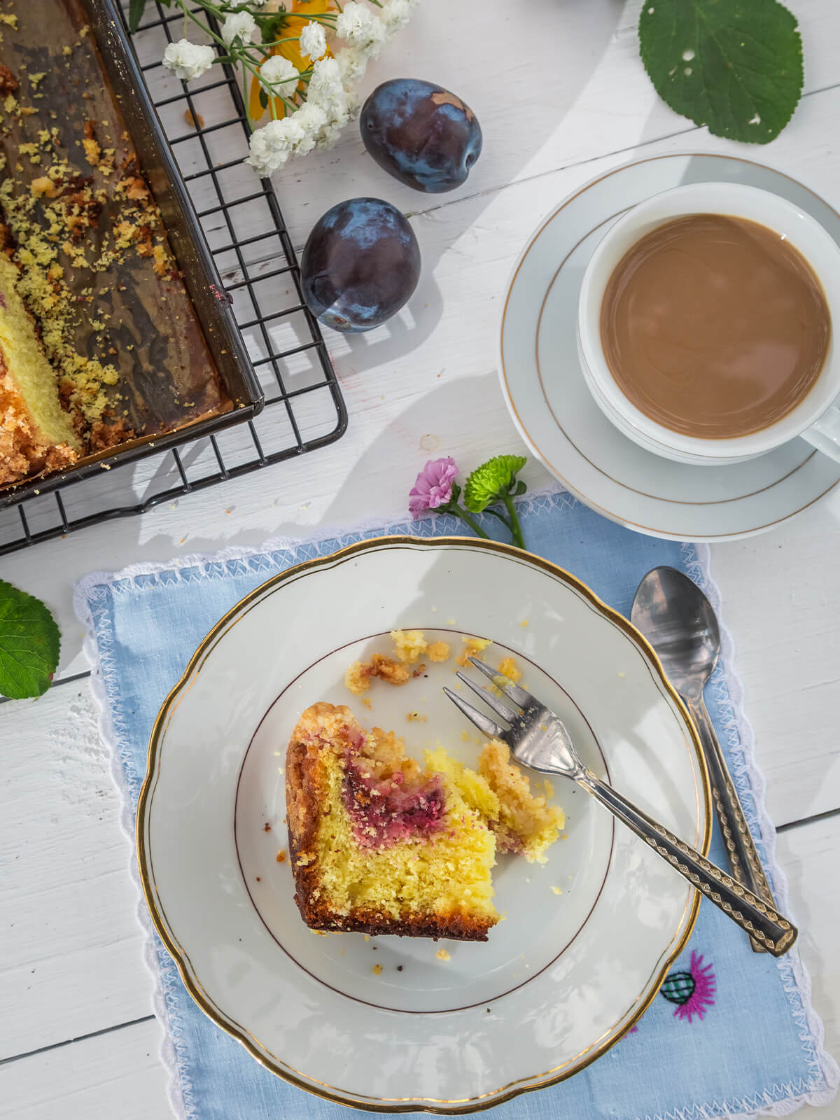 Stück Pflaumen-Streuselkuchen auf einem Teller mit Kaffee im Garten, im Hintergrund der Kuchen auf einem Gitter