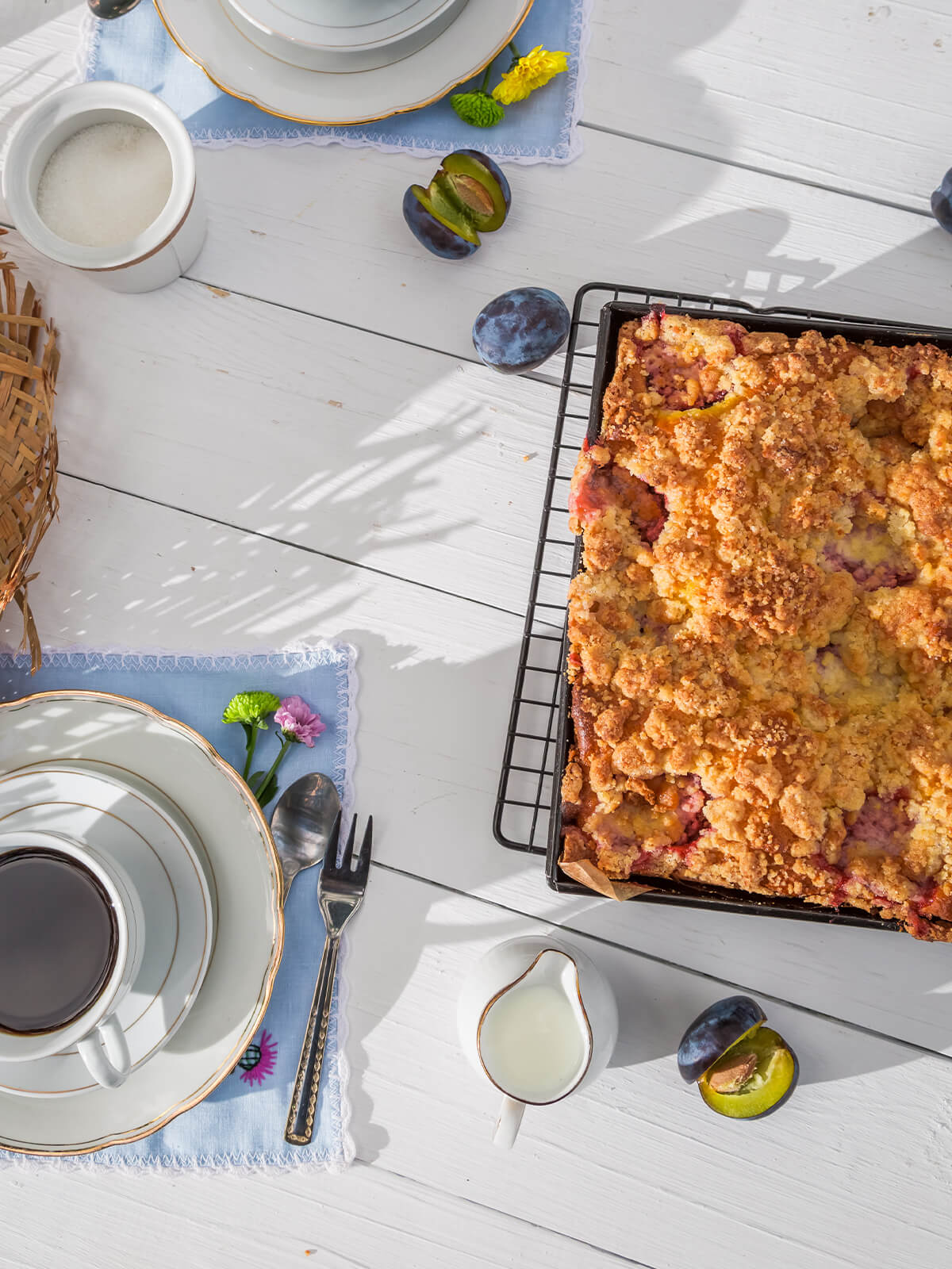 Pflaumen-Streuselkuchen in der hohen Form von oben, daneben eine Kaffeetasse und frische Pflaumen auf hellem Holz