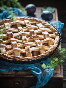 Fertige, goldbraun gebackene Pflaumen-Tarte mit Mürbeteig-Gitter und gedrehter Kordel, mit Puderzucker bestäubt, schräg von der Seite fotografiert