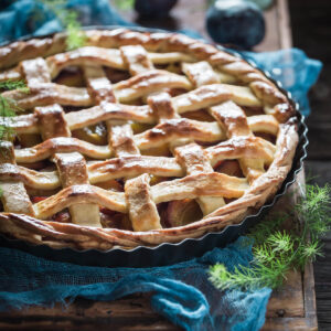 Fertige, goldbraun gebackene Pflaumen-Tarte mit Mürbeteig-Gitter und gedrehter Kordel, mit Puderzucker bestäubt, schräg von der Seite fotografiert