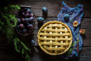 Ungebackene Pflaumen-Tarte mit von Hand geflochtenem hellem Mürbeteig-Gitter und gedrehter Teigkordel als Rand, daneben ein Korb dunkler Pflaumen