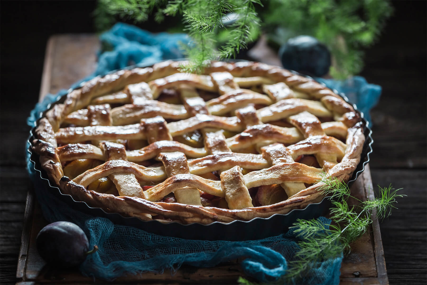 Goldbraun gebackene Pflaumen-Tarte mit glänzendem Mürbeteig-Gitter und gedrehter Kordel in der dunklen Tarteform, seitlich fotografiert