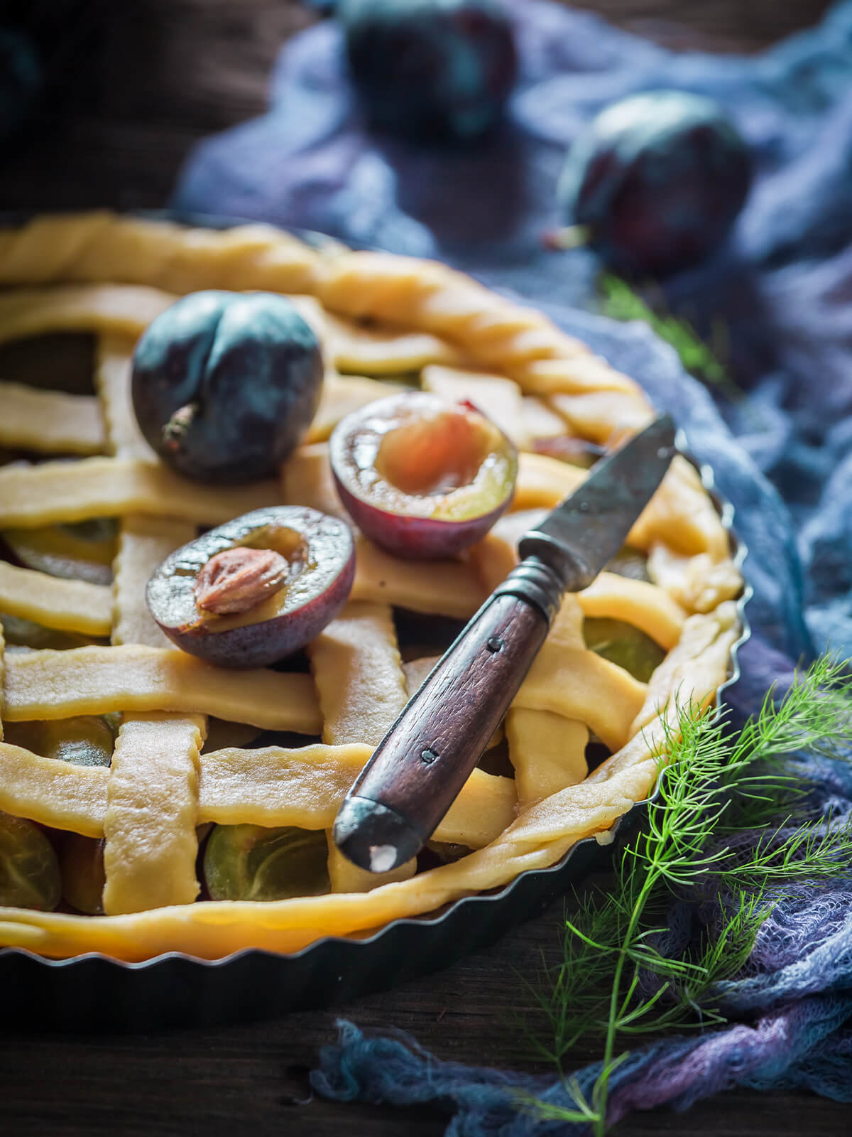 Nahaufnahme der ungebackenen Pflaumen-Tarte mit hellem Mürbeteig-Gitter, halbierten runden Pflaumen mit Stein und einem kleinen Messer