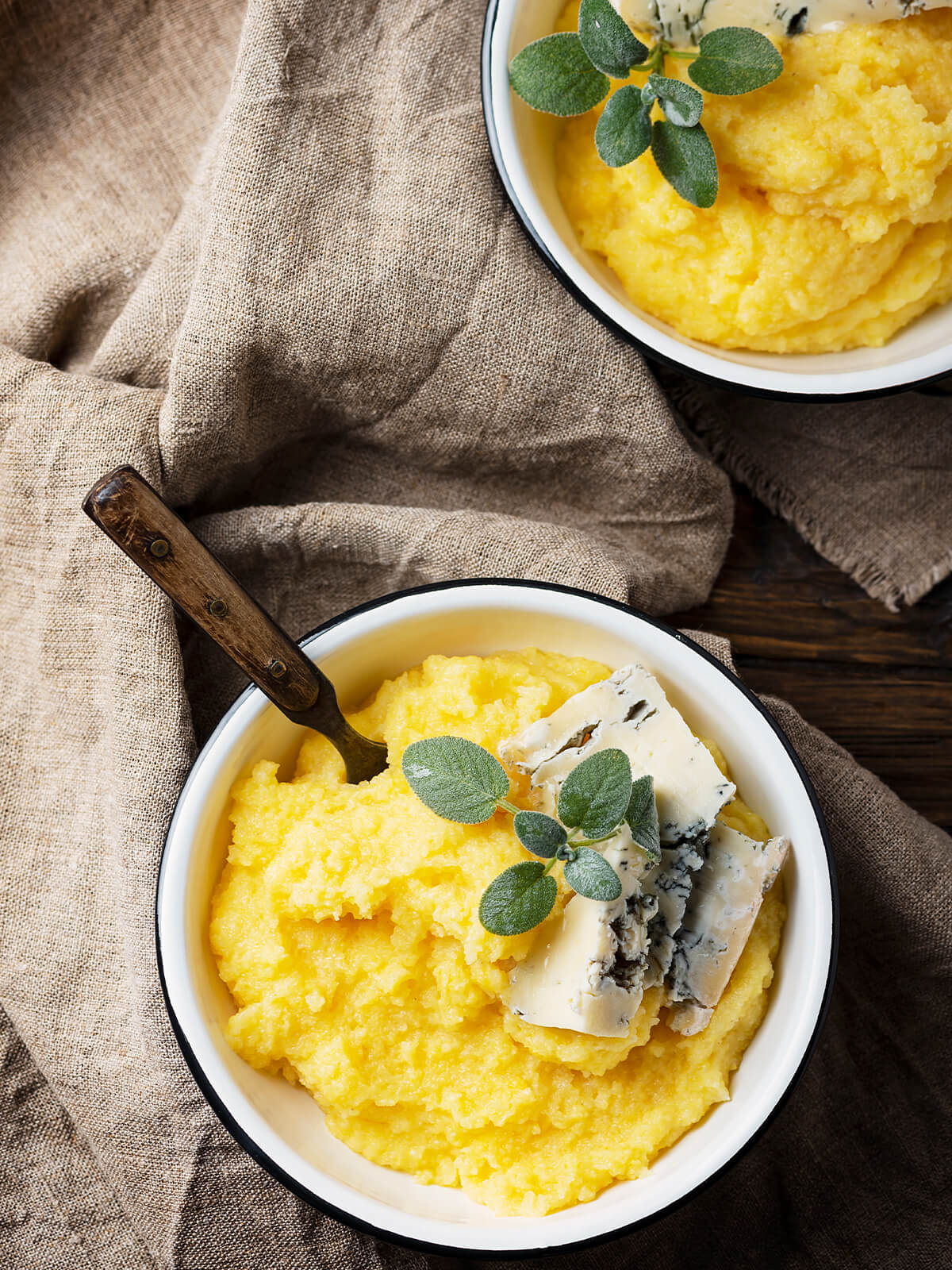 Zwei Schüsseln cremige Polenta mit Gorgonzola, frischem Salbei und Oregano, im Vordergrund eine Schüssel mit Löffel