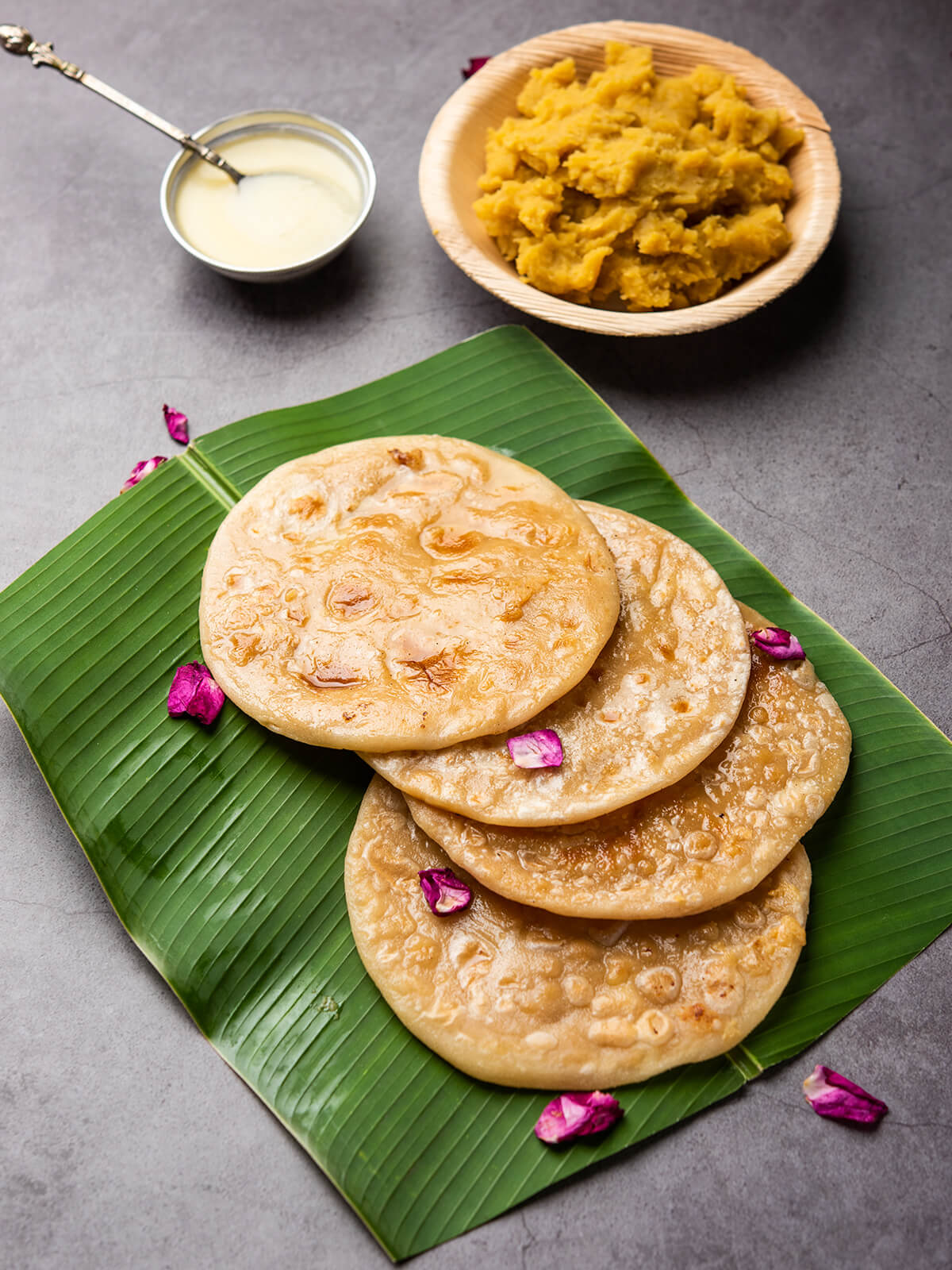 Vier goldbraune Puran Poli auf einem Bananenblatt, daneben eine Schale mit süßer Dal-Jaggery-Füllung und Ghee, garniert mit Rosenblättern