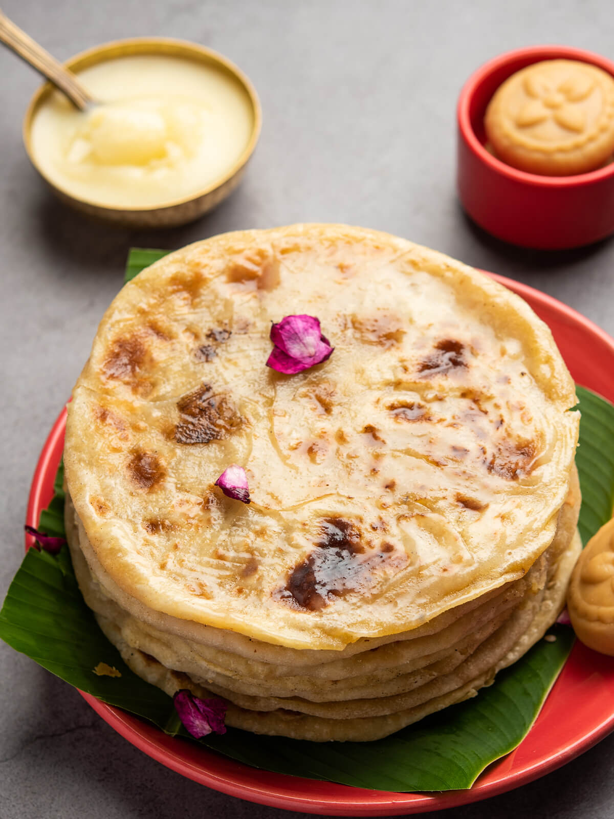 Ein Stapel goldbrauner Puran Poli auf einem roten Teller mit Bananenblatt, dazu eine Schale Ghee und Peda, ein indisches Milchkonfekt