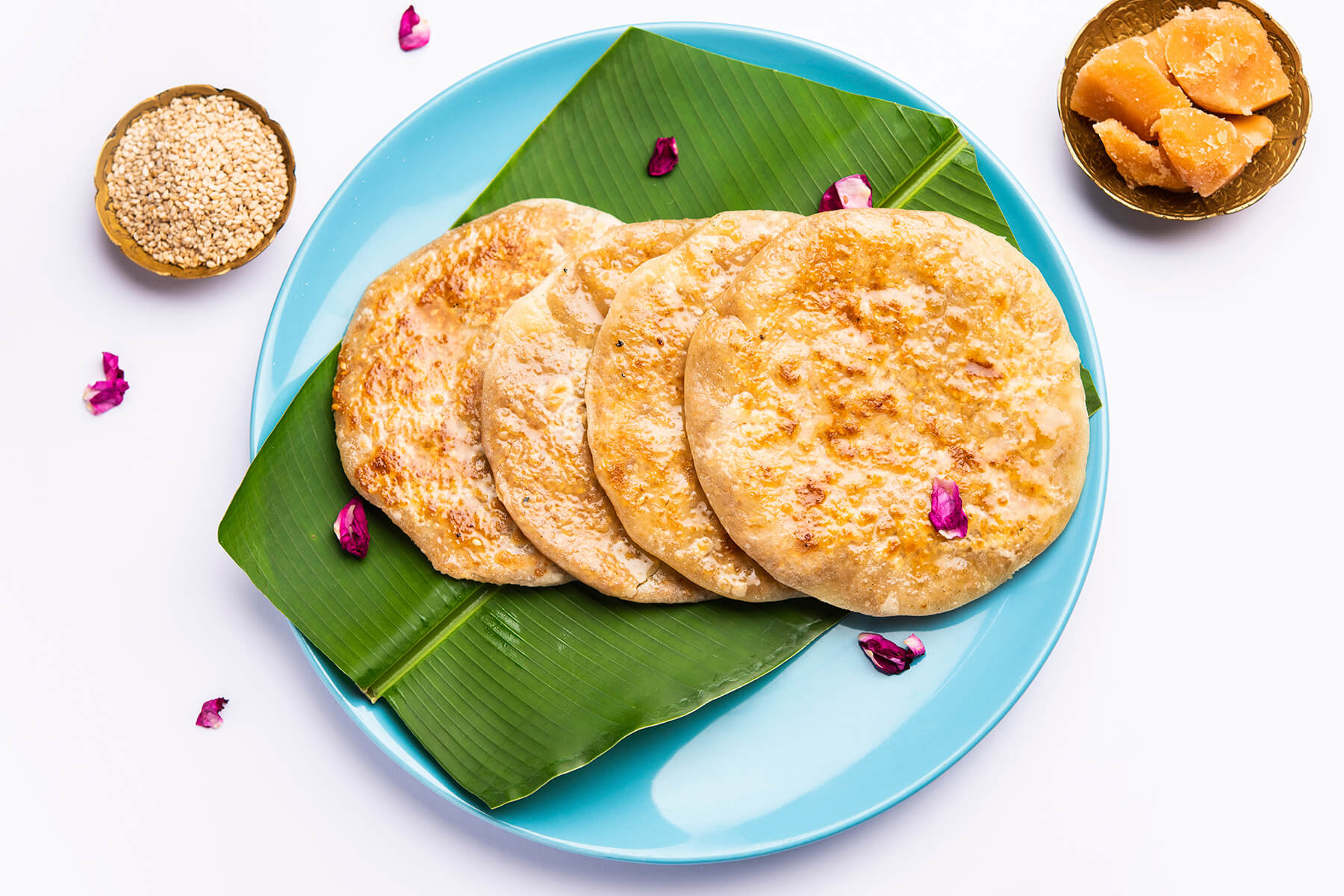 Vier Puran Poli auf einem türkisfarbenen Teller mit Bananenblatt, daneben Schalen mit Sesam und Jaggery