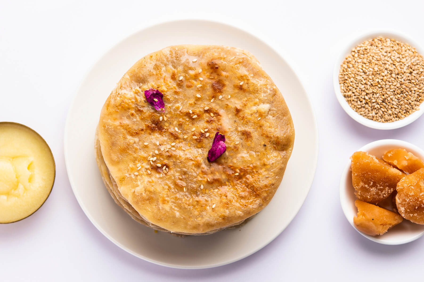 Puran Poli von oben auf einem hellen Teller, bestreut mit Sesam und Rosenblättern, daneben Ghee, Sesam und Jaggery
