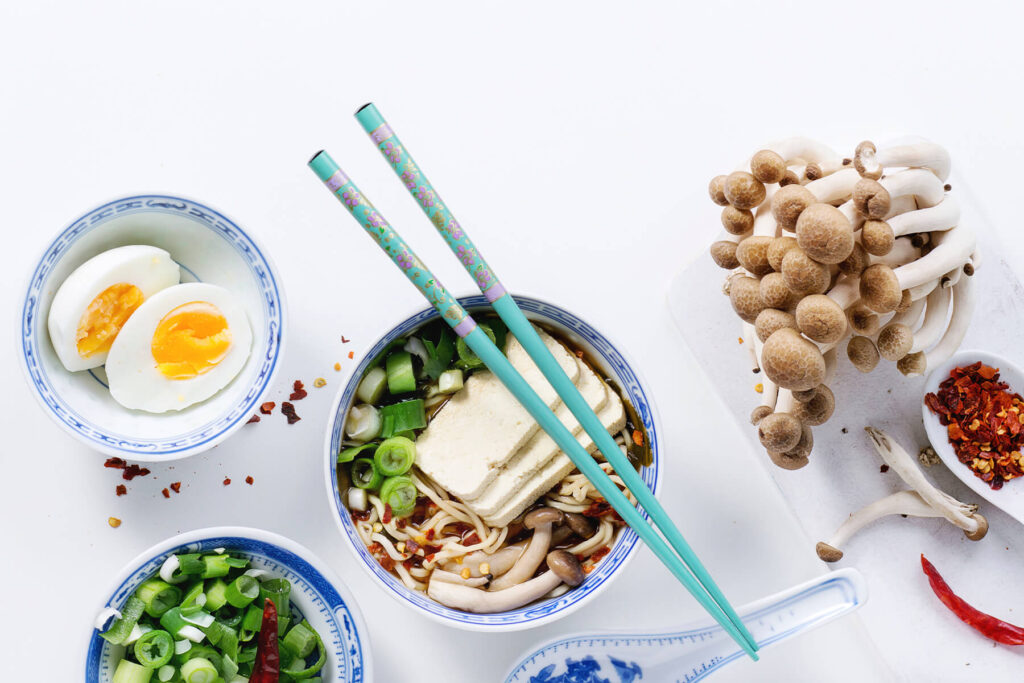 Vegetarische Ramen-Suppe mit Tofu, Pilzen, Nudeln und Frühlingszwiebeln von oben, daneben ein halbiertes gekochtes Ei, frische Buchenpilze und Chiliflocken
