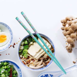 Vegetarische Ramen-Suppe mit Tofu, Pilzen, Nudeln und Frühlingszwiebeln von oben, daneben ein halbiertes gekochtes Ei, frische Buchenpilze und Chiliflocken
