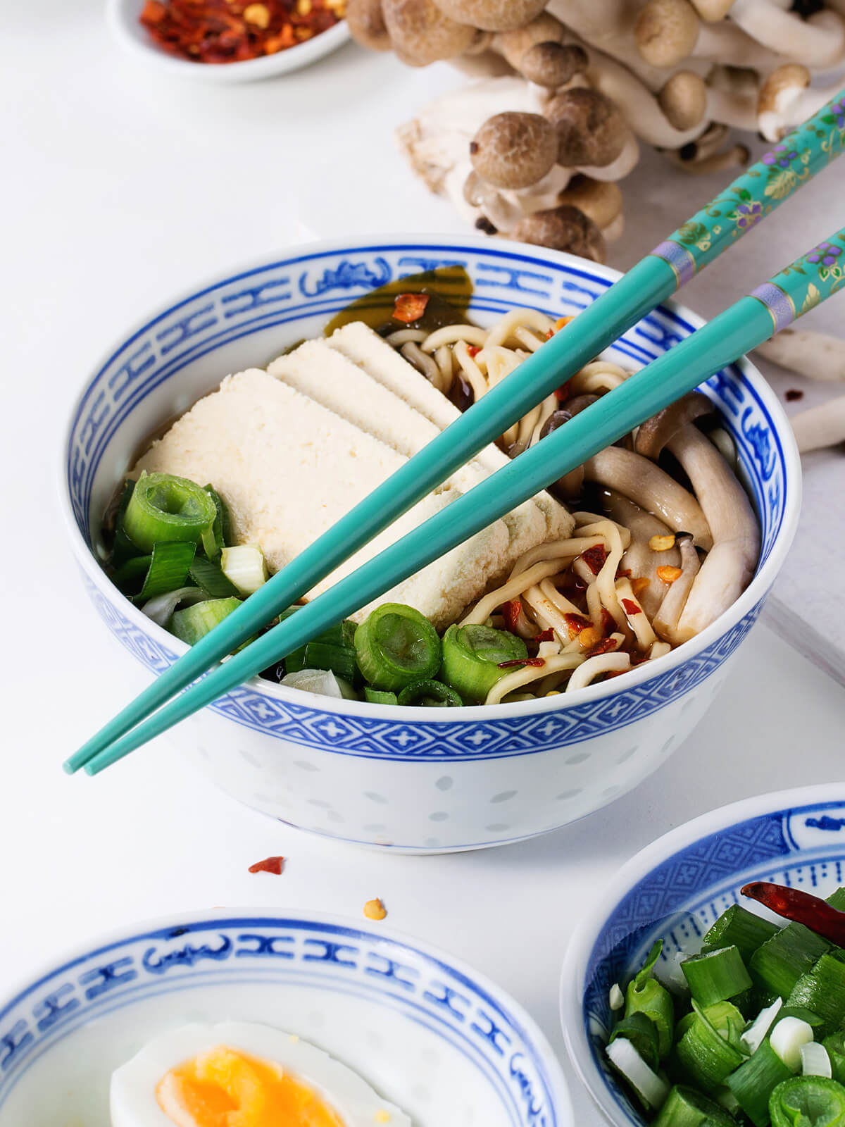 Ramen-Suppe mit Tofu und Pilzen in einer blau-weißen Schüssel, daneben frische Buchenpilze und ein halbiertes Ei
