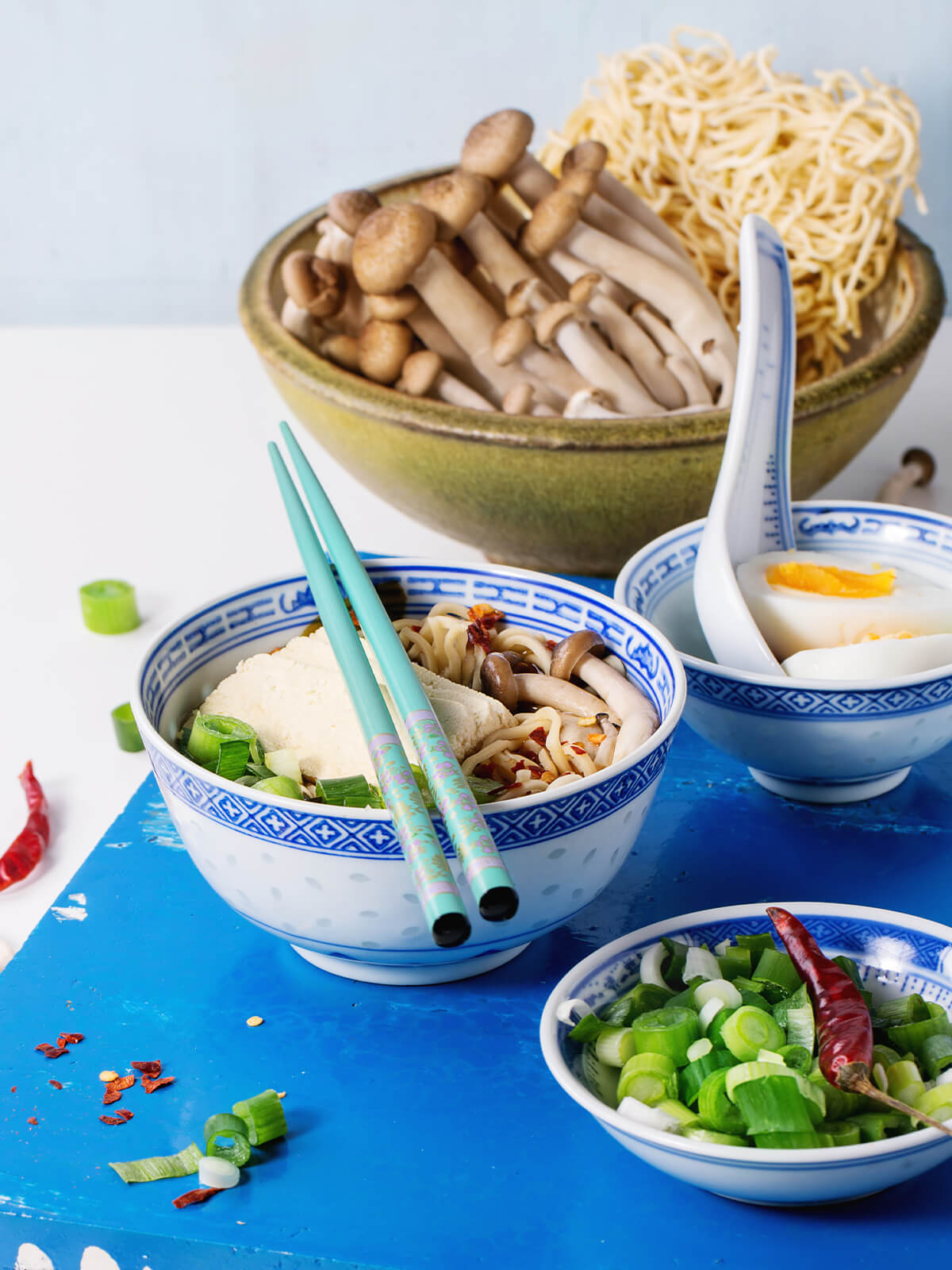 Angerichtete vegetarische Ramen-Suppe mit Tofu und Pilzen, daneben rohe Nudelnester, frische Buchenpilze, eine Schale Frühlingszwiebeln und ein halbiertes gekochtes Ei