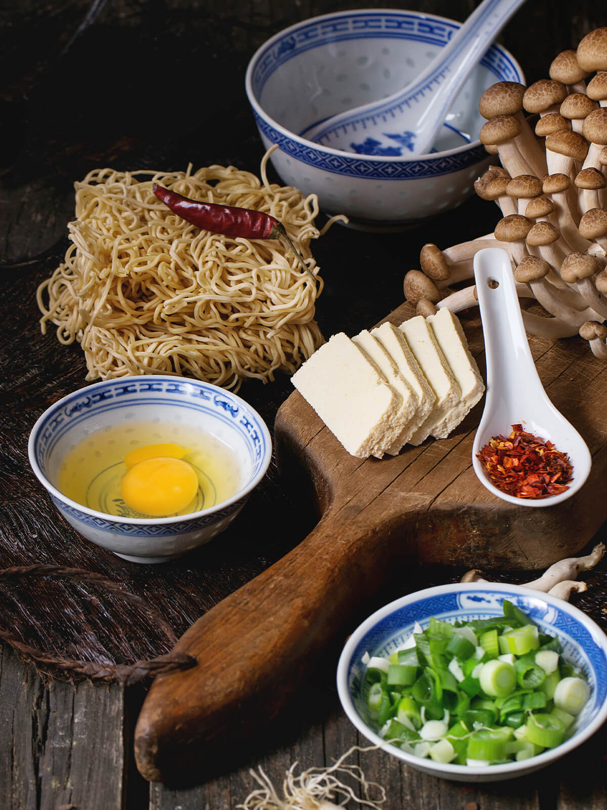 Zutaten für vegetarische Ramen-Suppe: Nudelnester, Tofu-Scheiben, Buchenpilze, zwei aufgeschlagene Eier, Chiliflocken und Frühlingszwiebeln