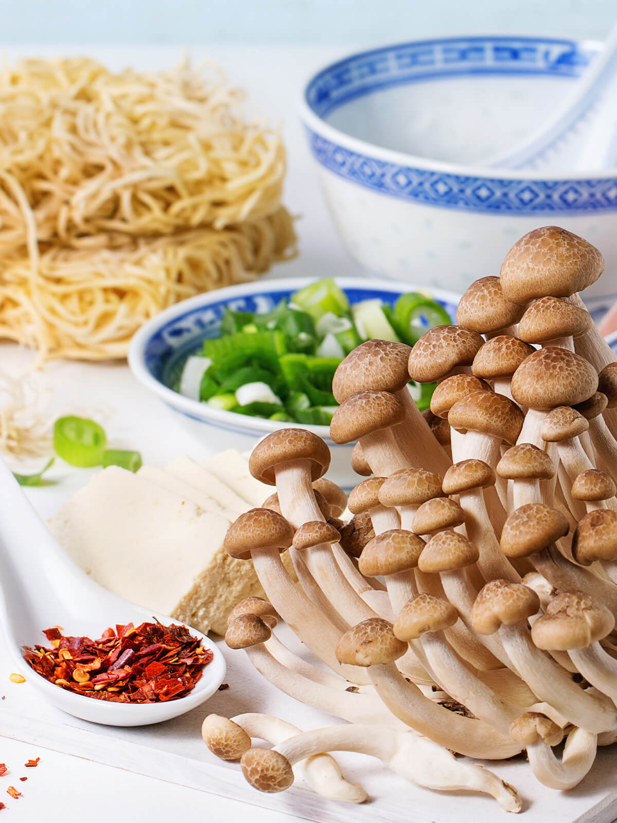 Zutaten für vegetarische Ramen-Suppe: Nudelnester, Buchenpilze, Tofu, Chiliflocken auf einem Löffel und eine Schale mit Frühlingszwiebeln