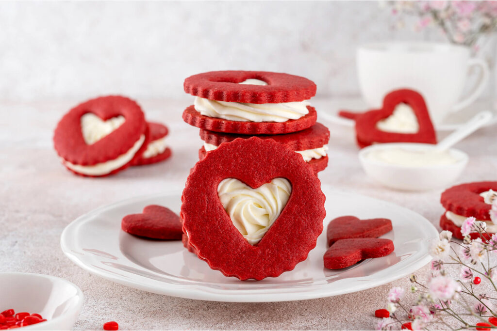 Aufrecht stehender Red Velvet Herz-Keks mit Frischkäse-Creme im Herzfenster auf einem weißen Teller, dahinter ein Stapel gefüllter Kekse, eine weiße Tasse und zarte rosa Blüten