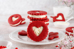 Aufrecht stehender Red Velvet Herz-Keks mit Frischkäse-Creme im Herzfenster auf einem weißen Teller, dahinter ein Stapel gefüllter Kekse, eine weiße Tasse und zarte rosa Blüten