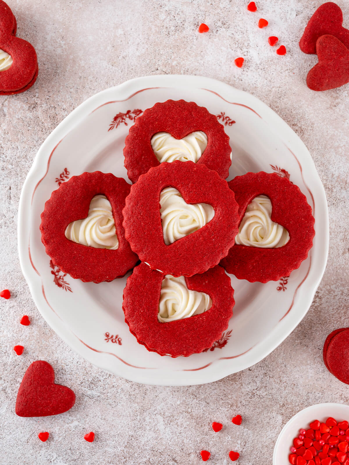 Fünf Red Velvet Herz-Kekse mit sichtbarer Frischkäse-Creme im Herzfenster, einer in der Mitte und vier kreuzförmig darum auf einem weißen Teller mit rotem Blumendekor