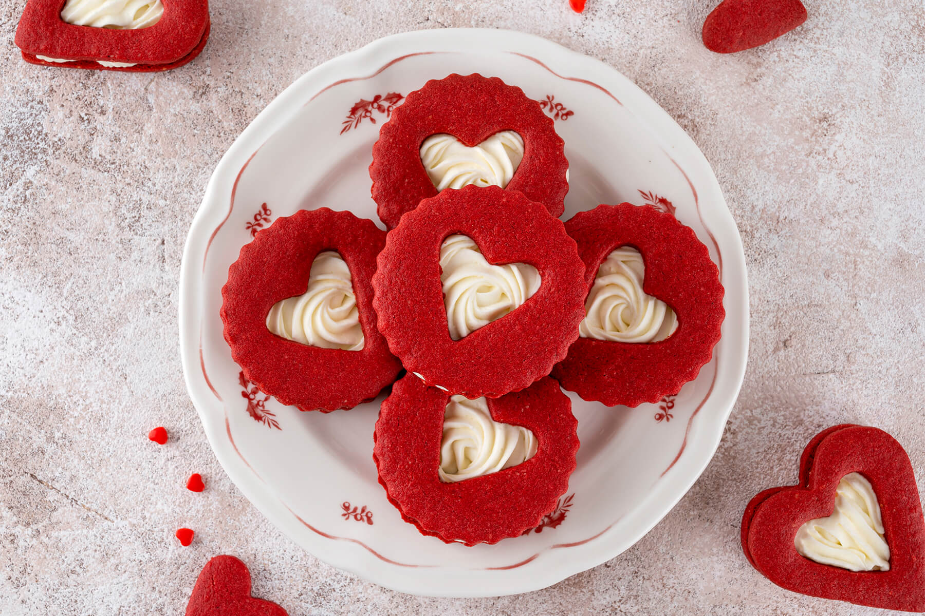 Fünf Red Velvet Herz-Kekse mit Frischkäse-Creme im Herzfenster, einer in der Mitte und vier kreuzförmig darum auf einem weißen Teller mit rotem Blumendekor, ringsum rote Zuckerherzen und herzförmige Kekse