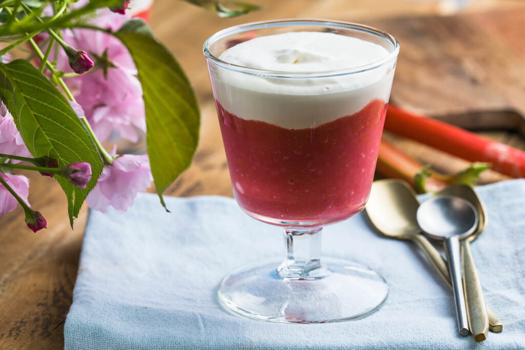 Rhabarber-Creme-Dessert im Glas mit roter Rhabarberschicht und weißer Vanille-Sahne-Creme, dekoriert mit Blüten