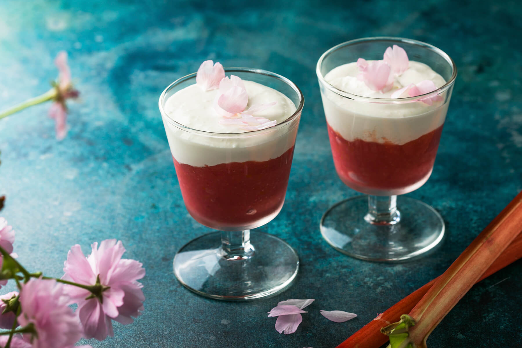 Zwei Gläser Rhabarber-Creme-Dessert mit roter Frucht- und weißer Cremeschicht, dekoriert mit Blütenblättern