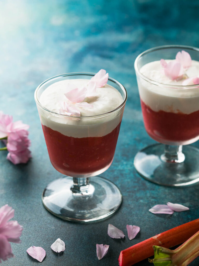 Zwei Gläser Rhabarber-Creme-Dessert mit roter Frucht- und weißer Cremeschicht, garniert mit Blüten