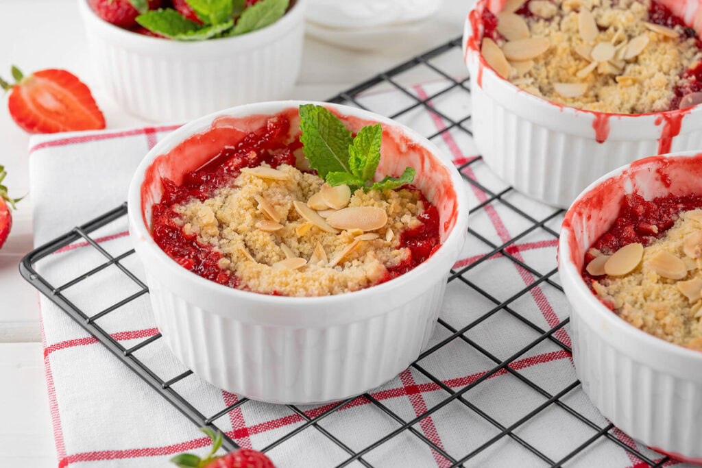 Mehrere Förmchen Rhabarber-Erdbeer-Crumble auf einem Abkühlgitter, mit leuchtend roter Frucht, goldenen Streuseln und Mandelblättchen, eines mit Minze garniert, daneben frische Erdbeeren