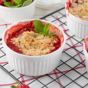Mehrere Förmchen Rhabarber-Erdbeer-Crumble auf einem Abkühlgitter, mit leuchtend roter Frucht, goldenen Streuseln und Mandelblättchen, eines mit Minze garniert, daneben frische Erdbeeren