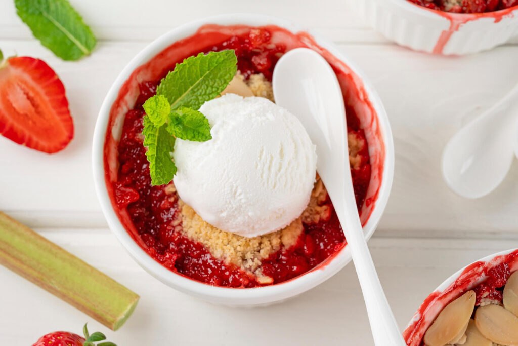Rhabarber-Erdbeer-Crumble in einem weißen Schälchen von oben, mit rotem Fruchtkompott, goldenen Streuseln, einer Kugel weißem Vanilleeis und einem Minzblatt, dazu ein Löffel