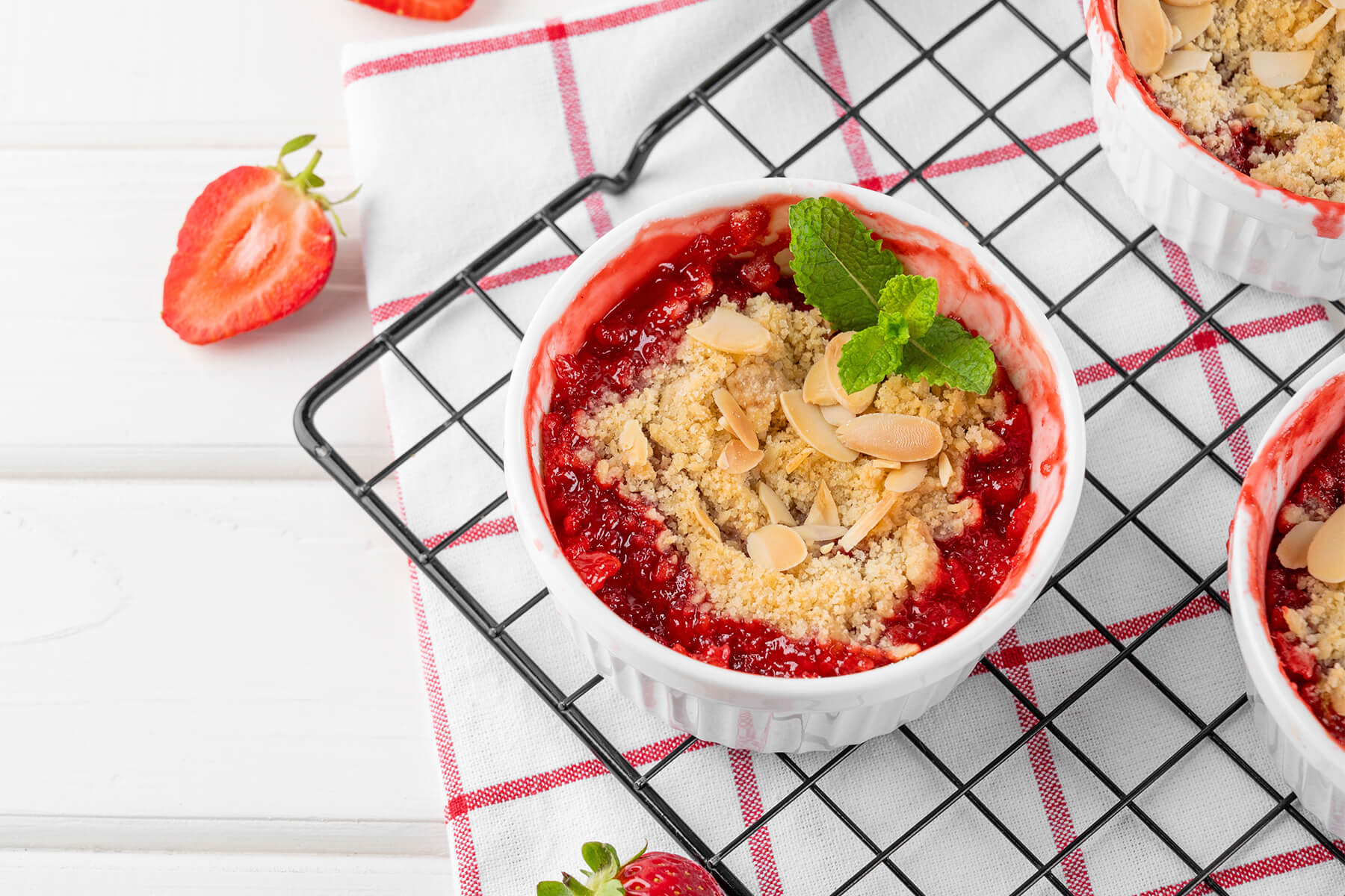 Ein Förmchen Rhabarber-Erdbeer-Crumble in Nahaufnahme auf einem Abkühlgitter, mit leuchtend rotem Fruchtkompott, goldenen Streuseln, Mandelblättchen und einem Minzblatt, daneben eine halbierte Erdbeere