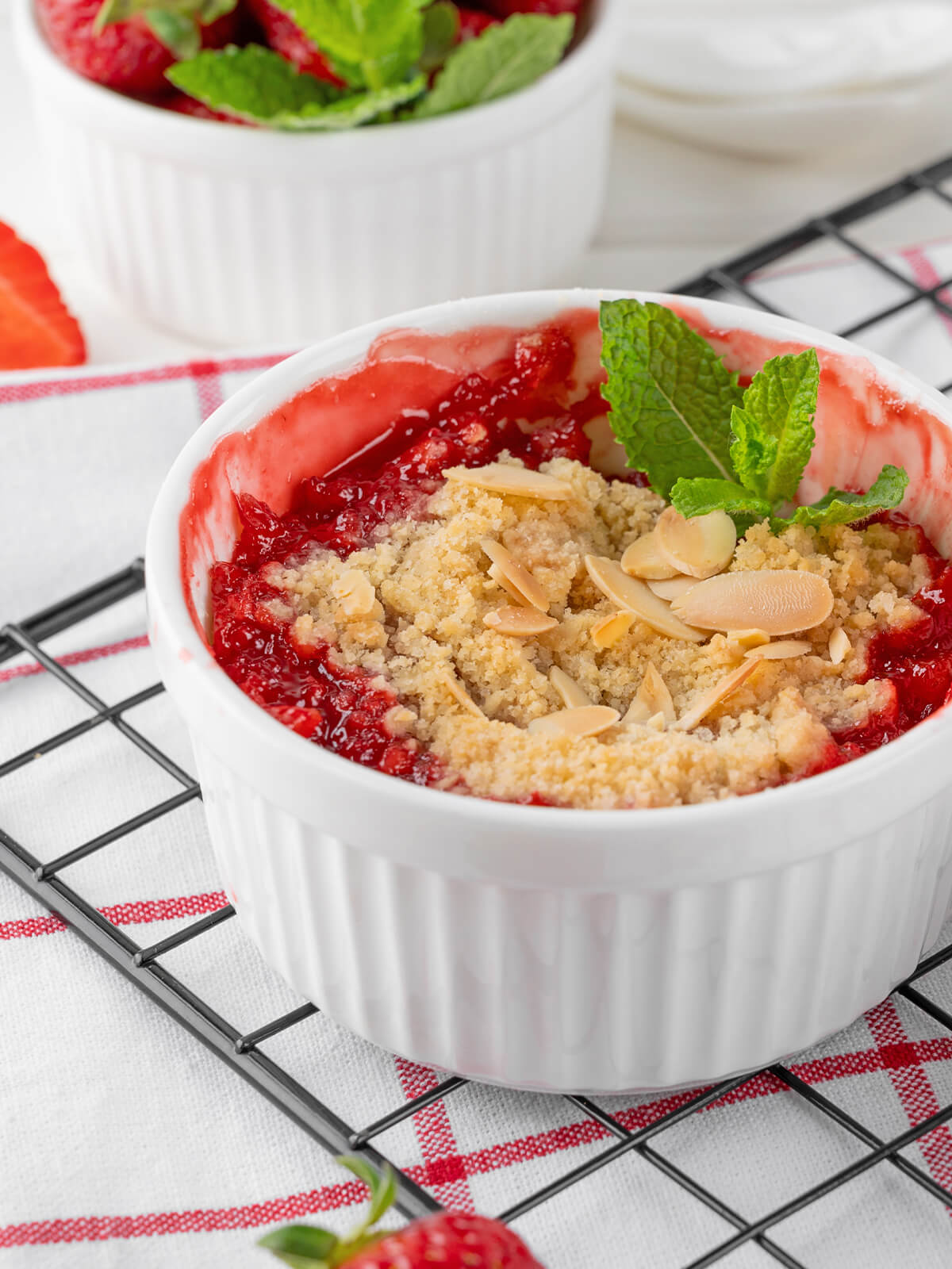 Ein Förmchen Rhabarber-Erdbeer-Crumble schräg von der Seite in Nahaufnahme auf einem Abkühlgitter, mit rotem Fruchtkompott, hell goldgelben Streuseln, Mandelblättchen und einem Minzblatt, daneben eine halbierte Erdbeere