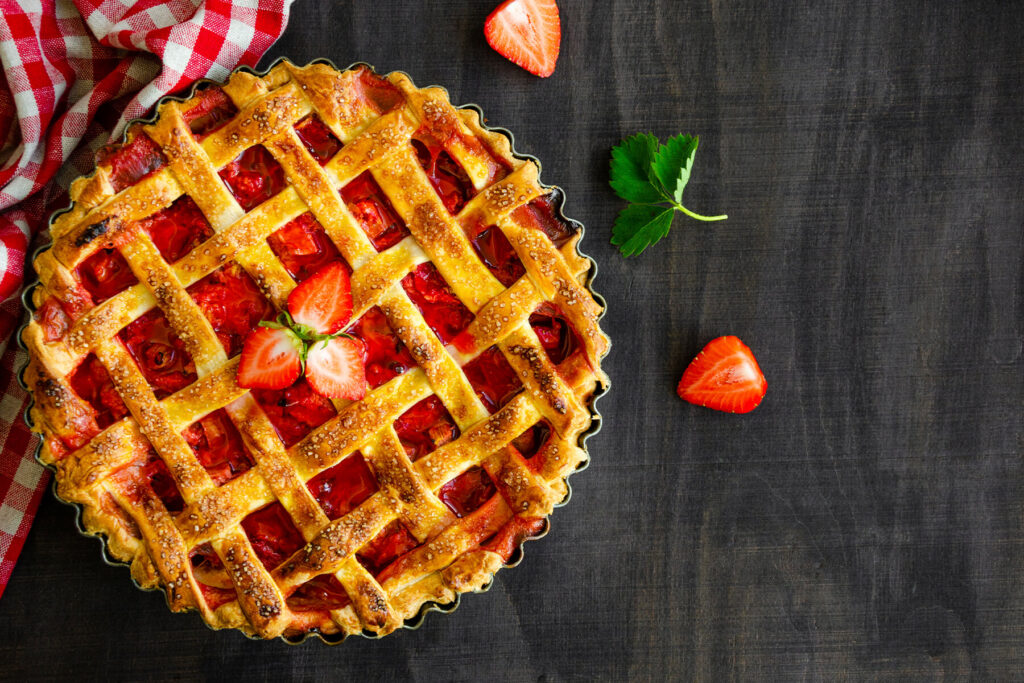 Ein ganzer Rhabarber-Erdbeer-Kuchen von oben in einer geriffelten Tarteform, mit goldbraunem Teiggitter aus quer aufgelegten Streifen, grobem Rohrzucker bestreut und roter Frucht, in der Mitte mit frischen Erdbeerscheiben garniert